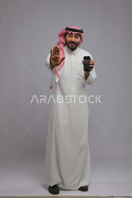Gestures to stop using modern technologies for useless things, displaying mobile phone applications and services, displaying a blank black screen on the mobile phone, portrait of a smiling Gulf Arab Saudi man wearing a shemagh and traditional thobe looking at the camera, full-length body photo, gray background