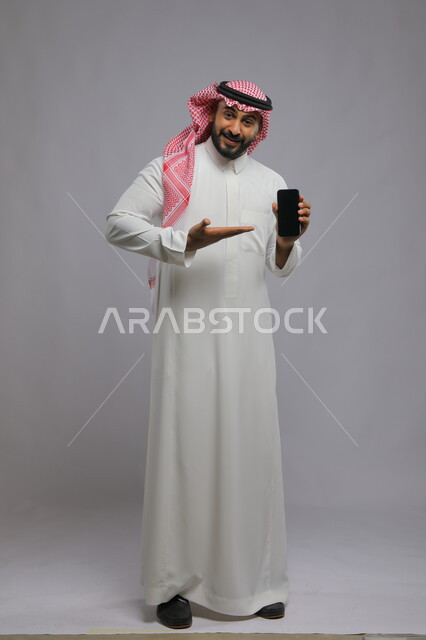 عرض تطبيقات وخدمات الهاتف المحمول ،الاشارة إلى الجوال بشاشة سوداء فارغة، استخدام التكنولوجيا المتطورة للتسويق الرقمي الالكتروني ، بورتريه لشاب عربي خليجي سعودي مبتسم يرتدي الشماغ و الثوب التقليدي ينظر إلى الكاميرا ،صورة الجسم بالطول الكامل، خلفية رمادية