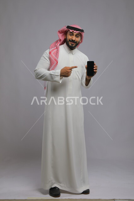 الاشارة إلى الجوال بشاشة سوداء فارغة، استخدام التكنولوجيا المتطورة للتسويق الرقمي الالكتروني ،عرض تطبيقات وخدمات الهاتف المحمول ، بورتريه لشاب عربي خليجي سعودي مبتسم يرتدي الشماغ و الثوب التقليدي ينظر إلى الكاميرا ،صورة الجسم بالطول الكامل، خلفية رمادية