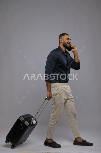  التواصل مع الأهل والأصدقاء, إجراء مكالمات هاتفية عبر الجوال, بورتريه لشاب عربي سعودي خليجي مبتسم يرتدي الزي الكاجوال يمسك بيده الهاتف المحمول وحقيبة السفر, حجز مباشر عبر تطبيقات وبرامج السياحة, صورة الجسم بالطول الكامل,  خلفية رمادية.