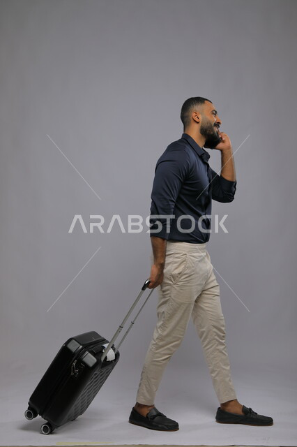 إجراء مكالمات هاتفية عبر الجوال, التواصل مع الأهل والأصدقاء, بورتريه لشاب عربي سعودي خليجي مبتسم يرتدي الزي الكاجوال يمسك بيده الهاتف المحمول وحقيبة السفر, حجز مباشر عبر تطبيقات وبرامج السياحة, صورة الجسم بالطول الكامل,  خلفية رمادية.