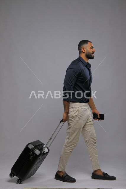 الشعور بالحماس والسعادة لقضاء اوقات ممتعة في الإجازة, تقنيات وأجهزة متطورة, بورتريه لشاب عربي سعودي خليجي مبتسم يرتدي الزي الكاجوال يمسك بيده الهاتف المحمول وحقيبة السفر, حجز مباشر عبر تطبيقات وبرامج التواصل الجوال, صورة الجسم بالطول الكامل,  خلفية رمادية.