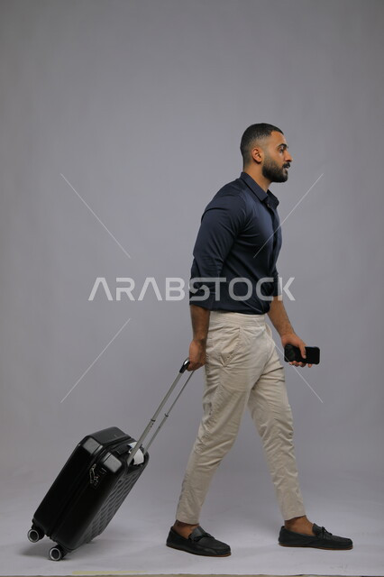 الاستعداد لقضاء رحلة صيفية ممتعة في الإجازة, تقنيات حديثة ومتطورة, بورتريه لرجل عربي سعودي خليجي يرتدي الزي الكاجوال يمسك بيده الهاتف المحمول وحقيبة السفر, حجز مباشر عبر تطبيقات وبرامج التواصل الجوال, صورة الجسم بالطول الكامل,  خلفية رمادية.