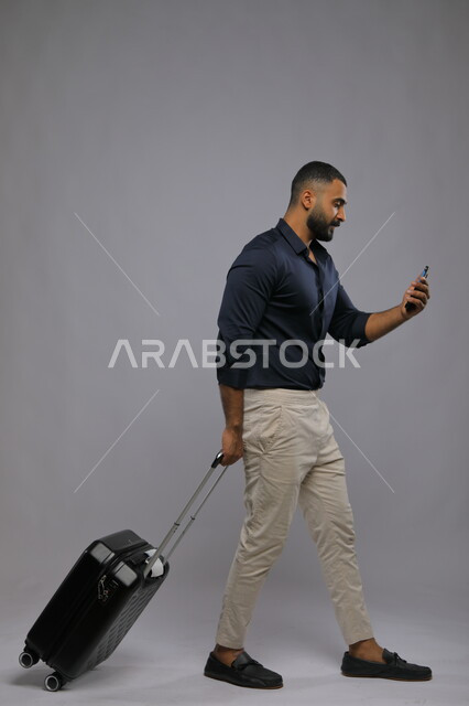 الاستعداد للمغادرة والسفر, قضاء عطلة صيفية ممتعة, بورتريه لرجل عربي سعودي خليجي مبتسم يرتدي الزي الكاجوال يمسك بيده الهاتف المحمول وحقيبة السفر, حجز مباشر عبر تطبيقات وبرامج التواصل الجوال, صورة الجسم بالطول الكامل,  خلفية رمادية.