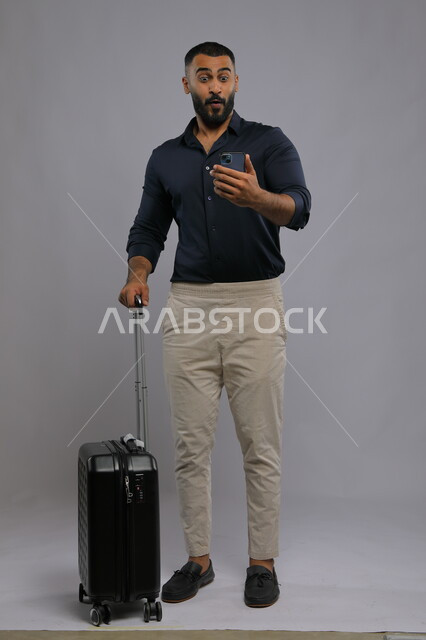 الشعور بالذهول والدهشة من ميزات التكنولوجيا المتطورة,  بورتريه لشاب عربي سعودي خليجي يرتدي الزي الكاجوال يمسك بيده الهاتف المحمول وحقيبة السفر, تصفح تطبيقات ومواقع السياحة عبر الجوال, صورة الجسم بالطول الكامل,  خلفية رمادية.
