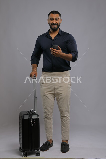 Direct booking via mobile applications and programs, enthusiastic gestures and looking at the camera, portrait of a smiling Saudi Arabian Gulf man wearing a casual outfit holding a mobile phone and a travel bag in his hand, use of advanced technology, full-length body portrait, gray background