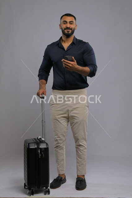Use of advanced technology, happy gestures and looking at the camera, portrait of a smiling Saudi Arabian Gulf man wearing a casual outfit holding a mobile phone and a travel bag in his hand, direct booking through mobile communication applications and programs, full-length body photo, gray background