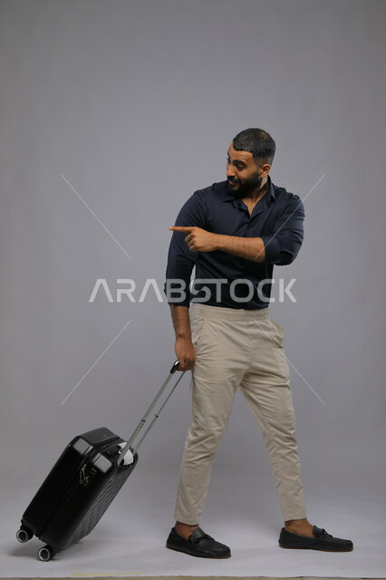 إيماءات السعادة والإشارة إلى شيء ما, السياحة في إجازة الصيف,  بورتريه لرجل عربي سعودي خليجي يرتدي الزي الكاجوال يجر حقيبة السفر, الاستعداد لقضاء رحلة صيفية ممتعة, صورة الجسم بالطول الكامل,  خلفية رمادية.