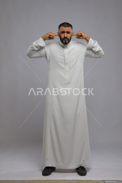 عدم تحمل الضوضاء والضجيج، بورتريه لرجل عربي سعودي خليجي يرتدي الزي التقليدي ويضع يداه على أذنيه، ايماءات وجه ويدين تدل على الانزعاج من الأصوات، الحفاظ على الهدوء في بيئة مزعجة، صورة الجسم بالطول الكامل، خلفية رمادية
