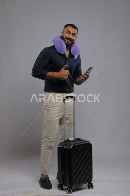 Thumbs up with the quality sign, portrait of a Saudi Gulf Arab man wearing casual clothes, using a sleeping pillow, holding a mobile phone and a travel bag, entertainment and entertainment during the trip using modern technologies, looking at the camera, full-length body photo, gray background