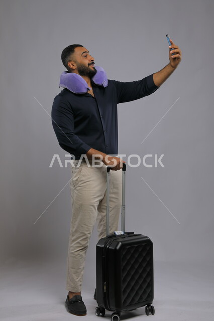 التقاط السيلفي لتوثيق اللحظات التذكارية أثناء الرحلة, مفهوم الاسترخاء والاستراحة, بورتريه لشاب عربي سعودي خليجي مبتسم يرتدي الزي الكاجوال يضع وسادة النوم على رقبته يمسك بيده الهاتف المحمول وحقيبة السفر, صورة الجسم بالطول الكامل,  خلفية رمادية.