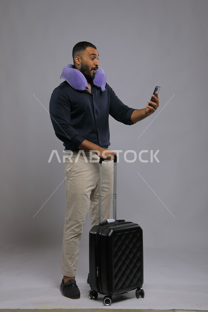 إجراء مكالمة فيديو مع الأهل والأصدقاء, إيماءات الذهول والدهشة, بورتريه لرجل عربي سعودي خليجي يرتدي الزي الكاجوال يضع وسادة الرقبة يمسك بيده الهاتف المحمول وحقيبة السفر, الحجز الالكتروني للسفر والسياحه عبر الجوال, صورة الجسم بالطول الكامل,  خلفية رمادية.