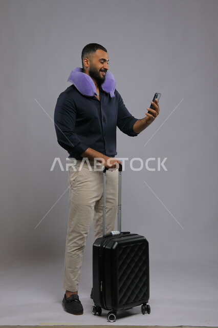 مشاهدة البرامج المفضلة عبر الجوال, مفهوم الاسترخاء والاستراحة, بورتريه لشاب عربي سعودي خليجي مبتسم يرتدي الزي الكاجوال يضع وسادة السفر يمسك بيده الهاتف المحمول وحقيبة السفر, قضاء أوقات ممتعه في السياحة بالعطلة الصيفيه, صورة الجسم بالطول الكامل,  خلفية رمادية.