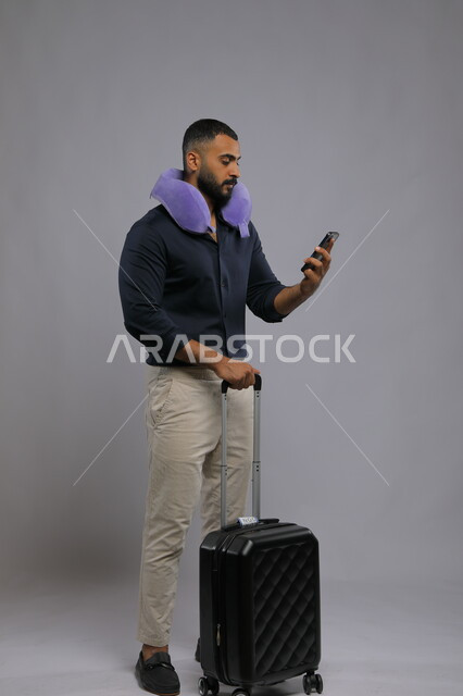 استخدام وسادة السفر للاسترخاء والاستراحة, بورتريه لرجل عربي سعودي خليجي يرتدي الزي الكاجوال يمسك بيده الهاتف المحمول وحقيبة السفر, قضاء أوقات ممتعه في السياحة بالعطلة الصيفيه, صورة الجسم بالطول الكامل,  خلفية رمادية.