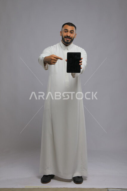 Using tablet programs and applications, portrait of a Saudi Gulf Arab man wearing traditional clothing and pointing to a blank black advertising screen on the tablet, browsing the Internet and completing tasks remotely, full body portrait, gray background
