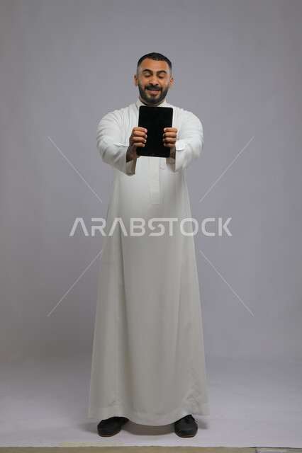 دور التقنية الحديثة في توفير راحة أكبر للأفراد، ايماءات الحماس والشغف باستخدام الجهاز اللوحي، بورتريه لرجل عربي سعودي خليجي يرتدي الزي التقليدي يمسك بيده التابلت بشاشه إعلانية للتسويق الالكتروني، صورة الجسم بالطول الكامل، خلفية رمادية