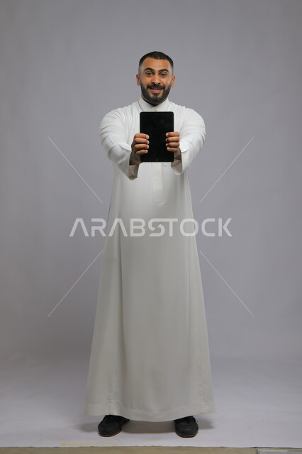 ايماءات الحماس والشغف باستخدام الجهاز اللوحي، بورتريه لرجل عربي سعودي خليجي يرتدي الزي التقليدي يمسك بيده التابلت بشاشه إعلانية للتسويق الالكتروني، دور التقنية الحديثة في توفير راحة أكبر للأفراد، صورة الجسم بالطول الكامل، خلفية رمادية