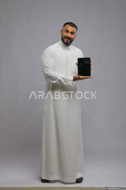 Using the tablet and modern technology to facilitate daily tasks, portrait of a Saudi Arabian Gulf man wearing traditional clothing holding a tablet in his hand with a blank black screen, the role of advanced technology in providing greater comfort for individuals, full-length body image, gray background