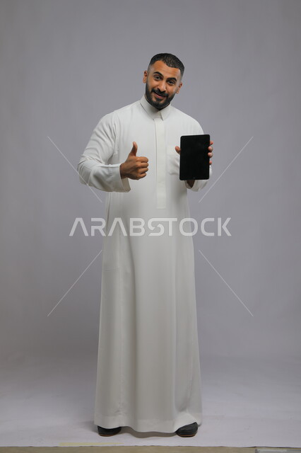 ايماءت يد تدل على الاعجاب بشيء ما، بورتريه لرجل عربي سعودي خليجي يرتدي الزي التقليدي يمسك بيده التابلت بشاشه سوداء فارغه، التسويق الإلكتروني وعرض خدمات وتطبيقات الجهاز اللوحي، صورة الجسم بالطول الكامل، خلفية رمادية