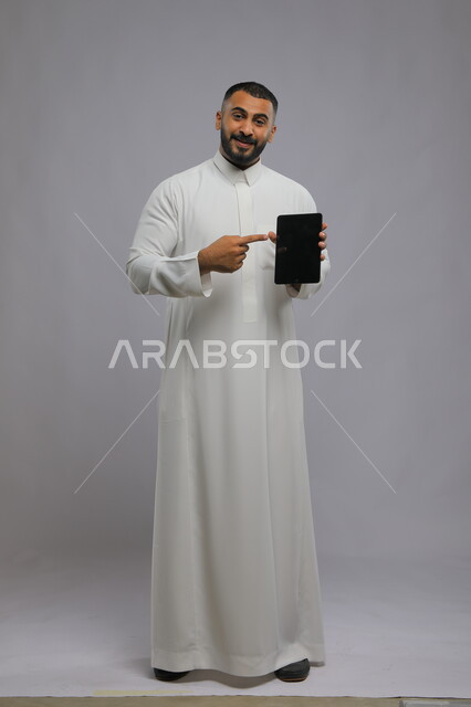 ايماءت الاعجاب باستخدام التقنيه والتكنولوجيا الحديثه، بورتريه لرجل عربي سعودي خليجي يرتدي الزي التقليدي ويشير بيده إلى التابلت بشاشه سوداء فارغه، التسويق الإلكتروني وعرض خدمات وتطبيقات الجهاز اللوحي،  صورة الجسم بالطول الكامل، خلفية رمادية