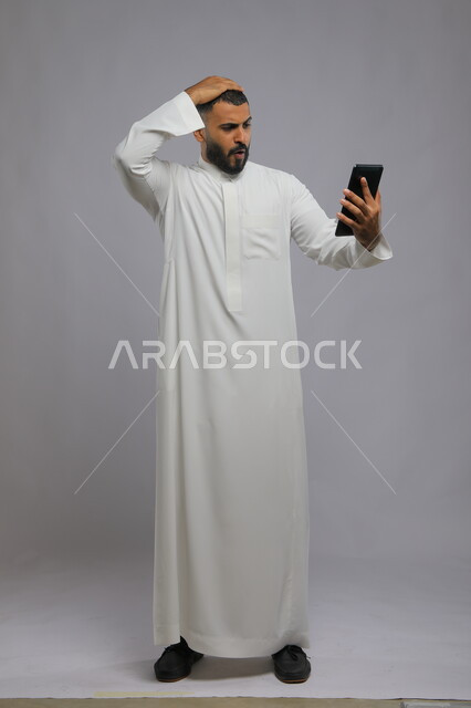 ايماءات الصدمة والدهشة، بورتريه لرجل عربي سعودي خليجي يرتدي الزي التقليدي ويمسك بيده التابلت، استعمال التقنيات الحديثة في التسويق الالكتروني، صورة الجسم بالطول الكامل، خلفية رمادية