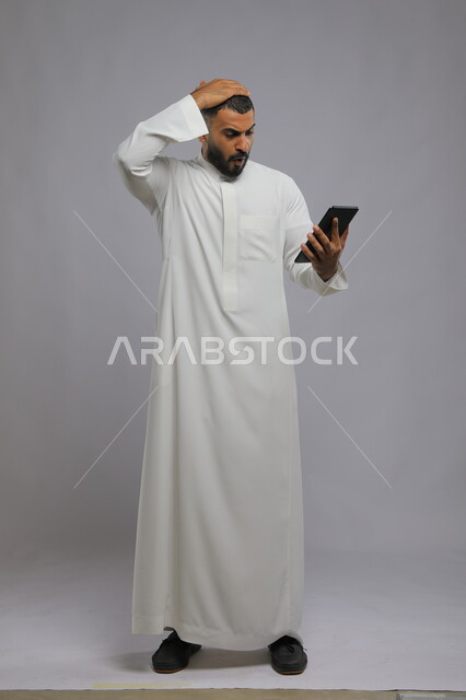 ايماءات الصدمة والدهشة، بورتريه لرجل عربي سعودي خليجي يرتدي الزي التقليدي ويمسك بيده التابلت، استعمال التقنيات الحديثة في التسويق الالكتروني، صورة الجسم بالطول الكامل، خلفية رمادية