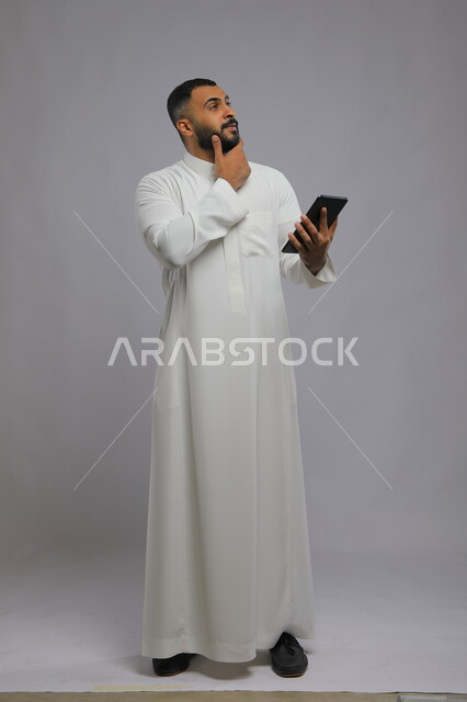 Gestures of thinking and contemplating digital content, portrait of a Saudi Gulf Arab man wearing traditional clothing and holding a tablet in his hand, the role of modern technology in providing greater comfort for individuals, full-length body image, gray background