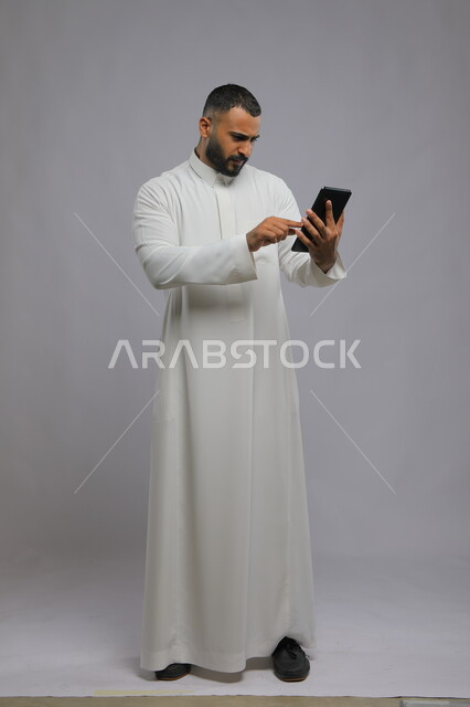 إستخدام برامج الجهاز اللوحي، بورتريه لرجل عربي سعودي خليجي يرتدي الزي التقليدي ويتواصل مع اصدقائه عبر التابلت، دمج التقنية والتكنولوجيا في الحياة العملية، صورة الجسم بالطول الكامل، خلفية رمادية