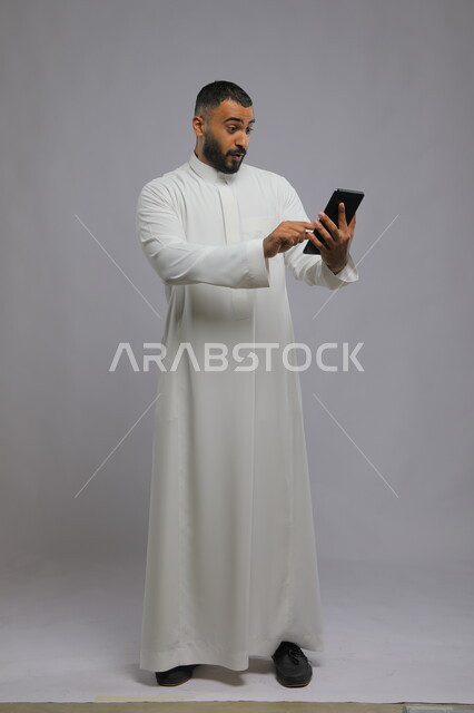 إستخدام برامج الجهاز اللوحي، بورتريه لرجل عربي سعودي خليجي يرتدي الزي التقليدي ويتواصل مع اصدقائه عبر التابلت، دمج التقنية والتكنولوجيا في الحياة العملية، صورة الجسم بالطول الكامل، خلفية رمادية