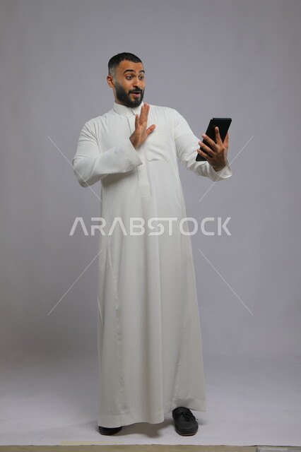 ايماءات الدهشة والتفاعل مع المحتوى الرقمي، بورتريه لرجل عربي سعودي خليجي يرتدي الزي التقليدي ويستخدم جهاز التابلت، دمج التقنية والتكنولوجيا في الحياة العملية، صورة الجسم بالطول الكامل، خلفية رمادية