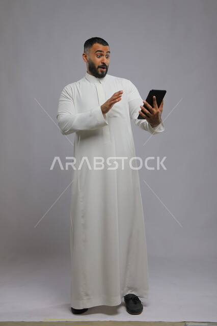 ايماءات الدهشة والتفاعل مع المحتوى الرقمي، بورتريه لرجل عربي سعودي خليجي يرتدي الزي التقليدي ويستخدم جهاز التابلت، دمج التقنية والتكنولوجيا في الحياة العملية، صورة الجسم بالطول الكامل، خلفية رمادية