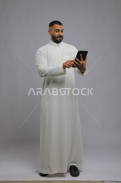 التسويق الإلكتروني وعرض خدمات وتطبيقات الجوال، بورتريه لرجل عربي سعودي خليجي يرتدي الزي التقليدي ويستخدم جهاز التابلت، ايماءت الاعجاب باستخدام التقنيه والتكنولوجيا الحديثه، صورة الجسم بالطول الكامل، خلفية رمادية