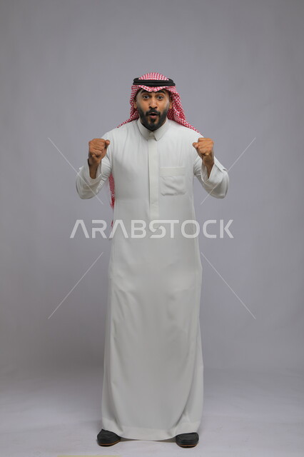 إيماءات الفوز والانتصار, بورتريه لرجل عربي خليجي سعودي مبتسم يرتدي الشماغ والثوب التقليدي يرفع يديه بتفاعل وحماس, الاندماج بتشجيع كرة القدم, صورة بالطول الكامل للجسم, خلفية رمادية