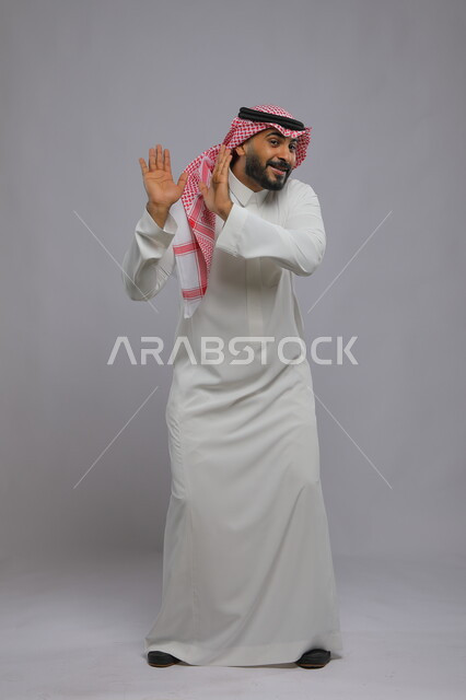حماية النفس من ضرر خارجي, رفع اليدين بإيماءات التوقف وعدم الاقتراب, بورتريه لرجل عربي خليجي سعودي يرتدي الثوب التقليدي والشماغ يقف بحذر, القلق والاحتياط والابتعاد عن شيء ما, صورة بالطول الكامل للجسم, خلفية رمادية