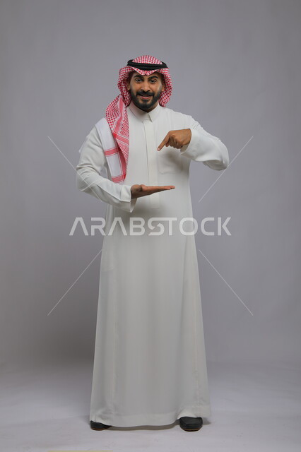 النظر إلى الكاميرا بإيماءات السرور, بورتريه لرجل عربي خليجي سعودي مبتسم يرتدي الثوب التقليدي والشماغ يفتح كفه للأعلى ويشير إلى عنصر ما, صورة الجسم بالطول الكامل, خلفية رمادية