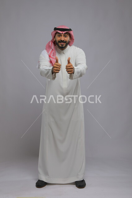 Standing straight and looking at the camera with gestures of interaction and approval, looks of enthusiasm and satisfaction, portrait of a Saudi Gulf Arab man wearing the traditional thobe and shemagh, raising his thumb up with the quality sign, full-length photo of the body, gray background