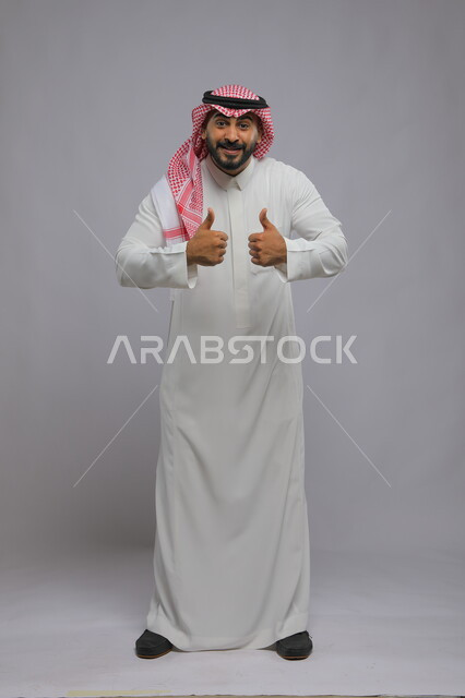 نظرات الحماس والشعور بالرضى, بورتريه لرجل عربي خليجي سعودي يرتدي الثوب والشماغ التقليدي يرفع إبهامه للأعلى بعلامة الجودة, الوقوف باستقامة والنظر إلى الكاميرا بإيماءات التفاعل والموافقة, صورة الجسم بالطول الكامل, خلفية رمادية