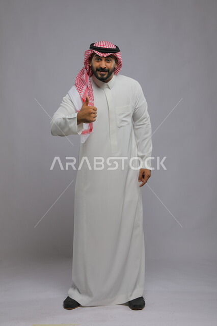 نظرات الحماس والشعور بالرضى, بورتريه لرجل عربي خليجي سعودي يرتدي الثوب والشماغ التقليدي يرفع إبهامه للأعلى بعلامة الجودة, الوقوف باستقامة والنظر إلى الكاميرا بإيماءات التفاعل والموافقة, صورة الجسم بالطول الكامل, خلفية رمادية