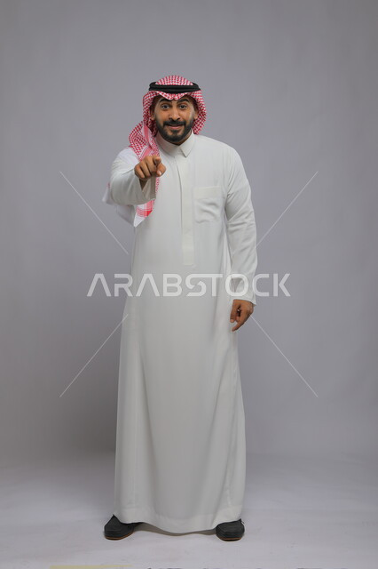 الاشارة إلى شيء ما بإيماءات الدهشة, تعابير التأكد والاستغراب, بورتريه لرجل عربي خليجي سعودي يرتدي الثوب التقليدي والشماغ يرفع إصبعه السبابة للتحديد والاختيار, صورة الجسم الكامل, خلفية رمادية