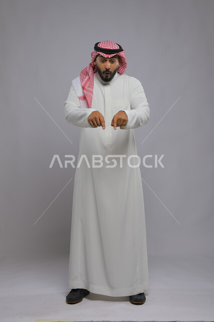 الوقوف باستقامة والإشارة إلى شيء ما, بورتريه لرجل عربي خليجي سعودي يرتدي الثوب التقليدي والشماغ يرفع إصابعه ويشير إلى هذا المكان بإيماءات الدهشة والحماس, صورة الجسم بالطول الكامل, خلفية رمادية