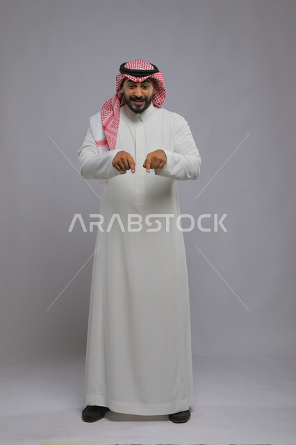 الوقوف باستقامة والإشارة إلى شيء ما, بورتريه لرجل عربي خليجي سعودي يرتدي الثوب التقليدي والشماغ يرفع إصابعه ويشير إلى هذا المكان بإيماءات الدهشة والحماس, صورة الجسم بالطول الكامل, خلفية رمادية