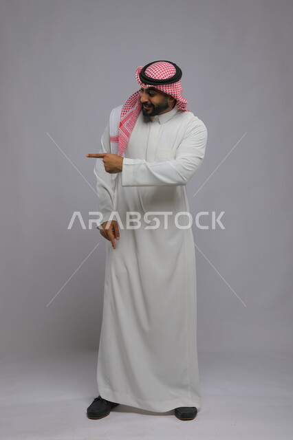 تعابير التأكد والتحديد, الاشارة إلى شيء ما بإيماءات الاكتشاف, بورتريه لرجل عربي خليجي سعودي يرتدي الثوب التقليدي والشماغ يرفع إصبعه السبابة للاختيار, صورة الجسم بالكامل, خلفية رمادية