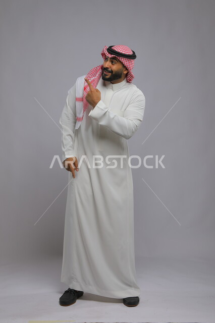 التعجب والاستغراب والدهشة من الأمر, الإشارة إلى شيء ما بإيماءات القلق, بورتريه لشاب عربي خليجي سعودي يرتدي الثوب التقليدي والشماغ يرفع إصبعه للأعلى لتحديد مكان ما, صورة لكامل الجسم, خلفية رمادية