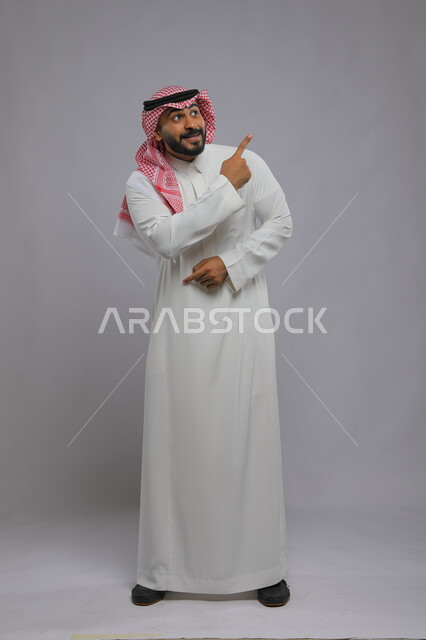 الخوف والاستغراب وعدم القبول والرضى, الإشارة إلى شيء ما بإيماءات القلق, بورتريه لشاب عربي خليجي سعودي يرتدي الثوب التقليدي والشماغ يرفع إصبعه للأعلى لتحديد, صورة الجسم بالطول الكامل, خلفية رمادية