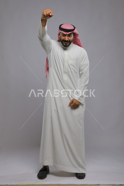 رفع اليد بإيماءات النصر والفوز, بورتريه لرجل عربي خليجي سعودي مبتسم يرتدي الثوب التقليدي والشماغ ينظر إلى الكاميرا بحماس, التفاعل والاندماج مع الألعاب الرياضية, صورة كامل الجسم, خلفية رمادية