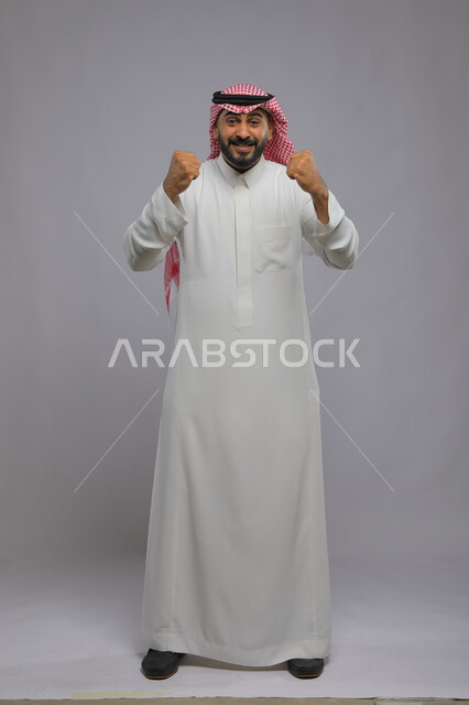 إيماءات الفوز والانتصار, بورتريه لرجل عربي خليجي سعودي مبتسم يرتدي الشماغ والثوب التقليدي يرفع يديه بتفاعل وحماس, الاندماج بتشجيع فريق كرة القدم, صورة الجسم بالكامل, خلفية رمادية