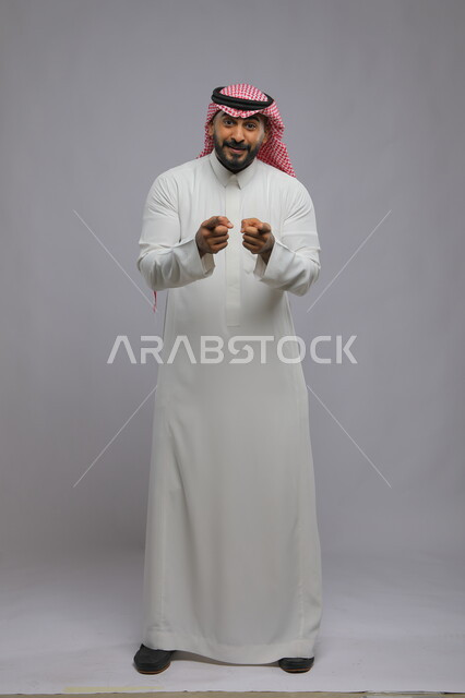 تعابير التأكد والاستغراب, الاشارة إلى شيء ما بإيماءات الدهشة, بورتريه لرجل عربي خليجي سعودي يرتدي الثوب التقليدي والشماغ يرفع أصابعه للتحديد والاختيار, صورة بطول الجسم الكامل, خلفية رمادية