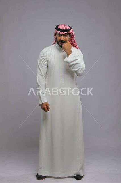 مفهوم تنفيذ الأوامر وتلبية الطلبات بالرحب والسعة, التأكيد على أمر معين, بورتريه لرجل عربي خليجي سعودي يرتدي الثوب التقليدي والشماغ يضع يديه على عينه, ايماءات التركيز والنظر لشيء ما, صورة الجسم بالطول الكامل, خلفية رمادية