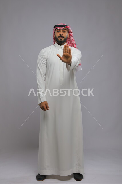 الصمت والتوقف عن الكلام, الأمر بالنهي عن فعل شيء ما, بورتريه لرجل عربي خليجي سعودي يرتدي الثوب والشماغ التقليدي يرفع كفه للأعلى, طلب خفض الصوت والتهديد, صورة لكامل الجسم, خلفية رمادية