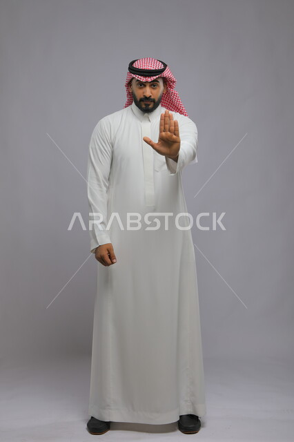 النهي عن فعل شيء ما, الصمت والتوقف عن الكلام, بورتريه لرجل عربي خليجي سعودي مغمض العينين يرتدي الثوب والشماغ التقليدي يرفع كفه للأعلى, طلب خفض الصوت والتهديد, صورة لكامل الجسم, خلفية رمادية
